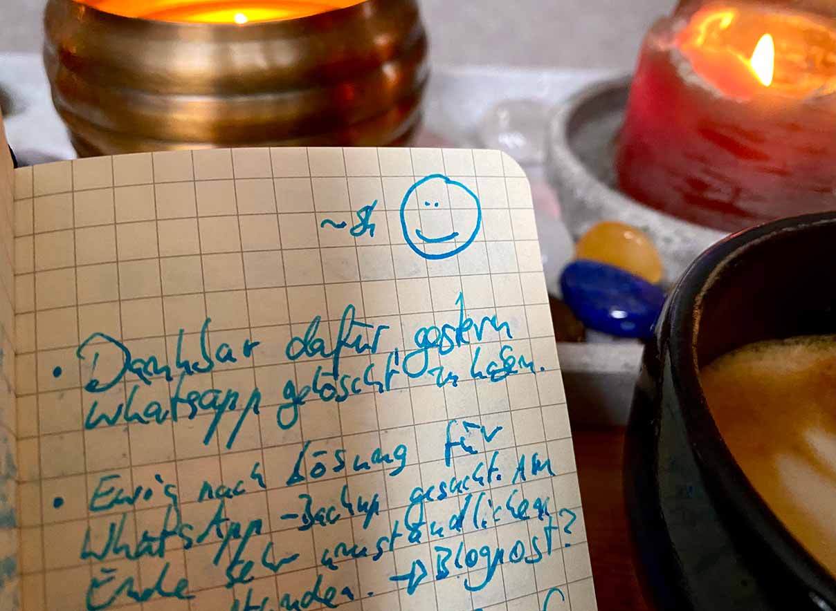 Kariertes Tagebuch wird neben einer Tasse Kaffee vor Tisch-Schmuck mit Kerzen und Edelsteinen hochgehalten. Die EInträge im Tagebuch erklären, dass WhatsApp endlich gelöscht ist.